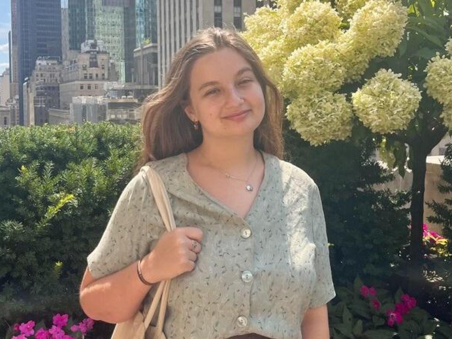 Tara Monastesse smiles, holding a tote bag over right shoulder with flowers and skyscrapers in the background