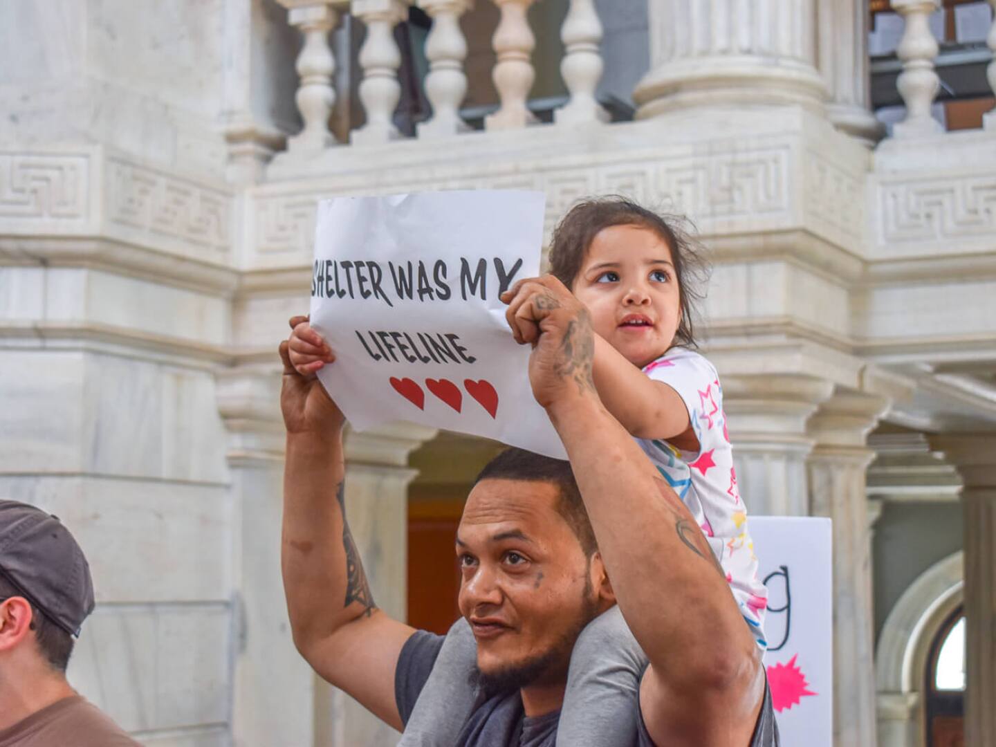 Mia Negron, 2, sits atop her father Luis’ shoulders at a State House rally on May 27, 2025. The family became unhoused when Mia was an infant in 2023.