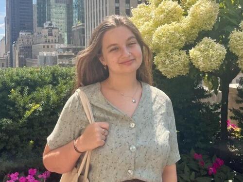 Tara Monastesse smiles, holding a tote bag over right shoulder with flowers and skyscrapers in the background